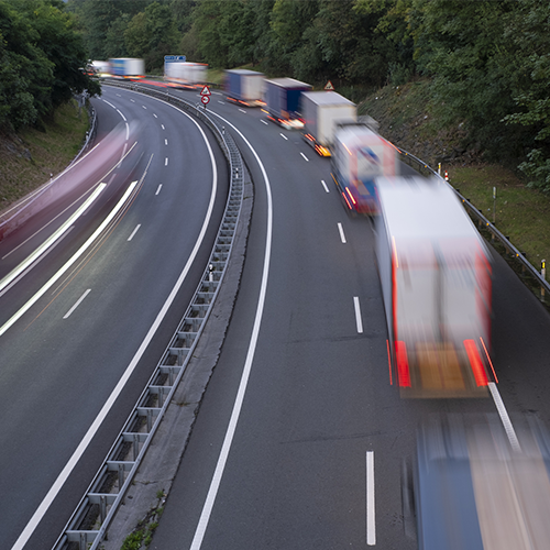 Multi-lane highway with several moving trucks, cars blurred by speed, curving road flanked by trees on both sides, daytime scene